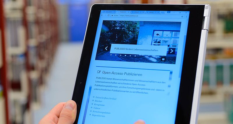 Eine Person, von der nur die Hand zu sehen ist, hält ein Tablet, auf dem die Website www.publisso.de zu sehen ist. Im Hintergrund gefüllte Regale in einer Bibliohek.